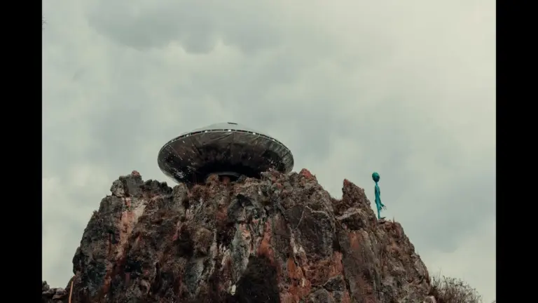 A UFO-shaped structure sits atop rugged rocks against a cloudy sky, with a small, green alien figure nearby, creating a mysterious, surreal atmosphere.