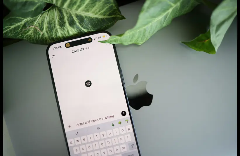 A smartphone showing a ChatGPT screen rests on a laptop with a visible Apple logo, surrounded by large green leaves, conveying a tech-nature theme.