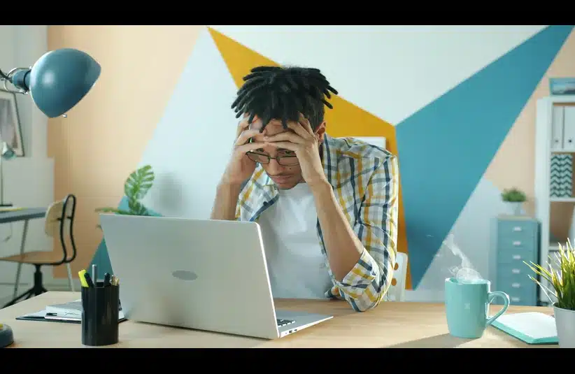 A person sitting at a desk in the workplace, looking stressed with their head in their hands, in front of a laptop. A steaming mug and office supplies are nearby. Overuse of AI is causing burnout.