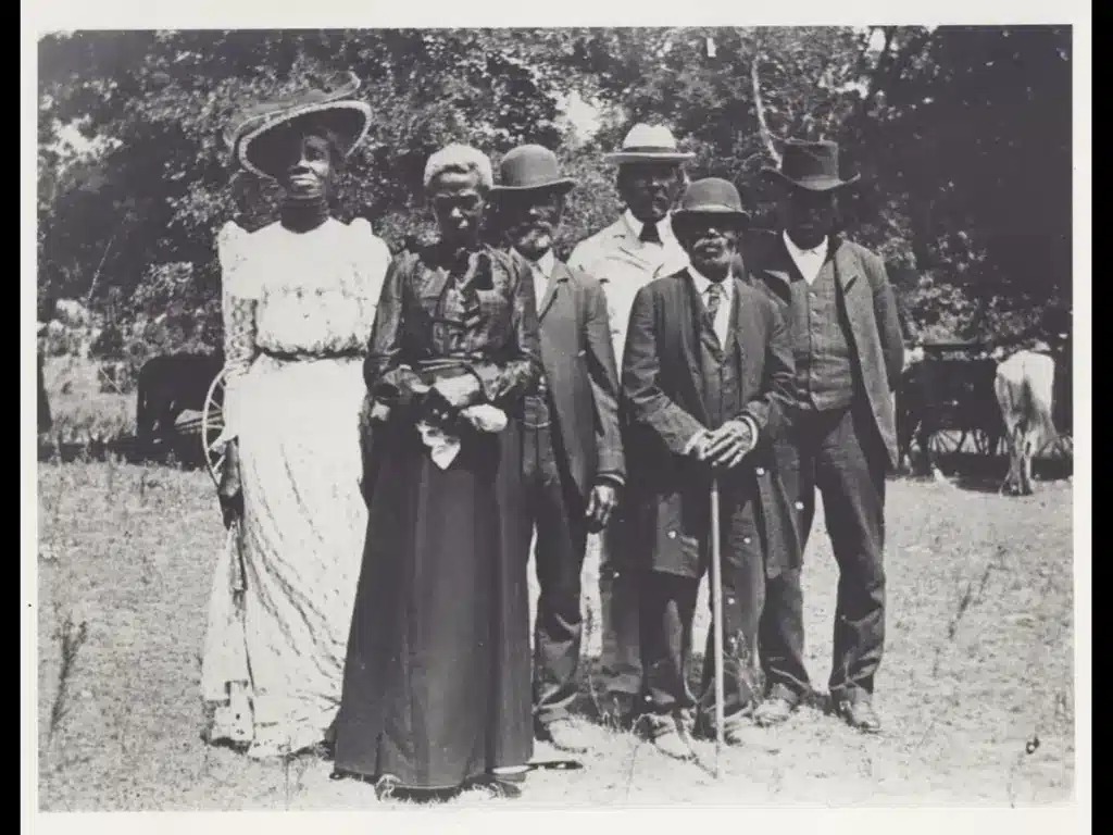 Historic black-and-white photo of six African American adults standing outdoors. They wear formal 19th-century attire, evoking a tone of dignity on Juneteenth in Texas.