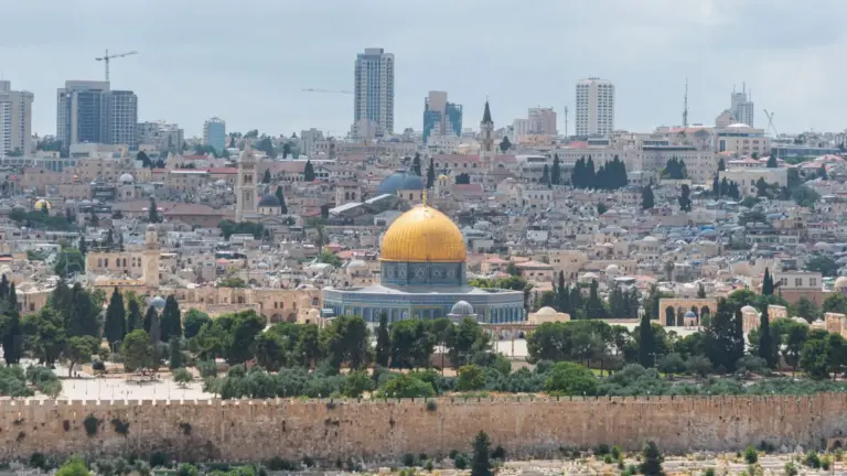 Panoramic view of Israel featuring the Dome of the Rock with its golden dome. The cityscape includes historic buildings, trees, and modern skyscrapers.