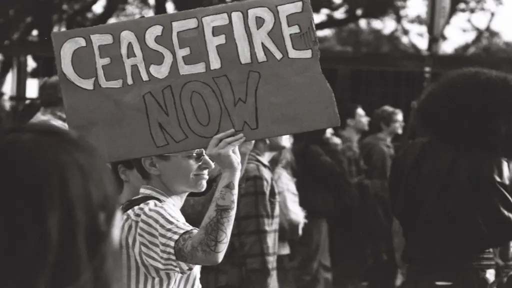 A person holds a "Ceasefire Now" sign at a protest. They're wearing glasses and a striped shirt, surrounded by others in a peaceful atmosphere.