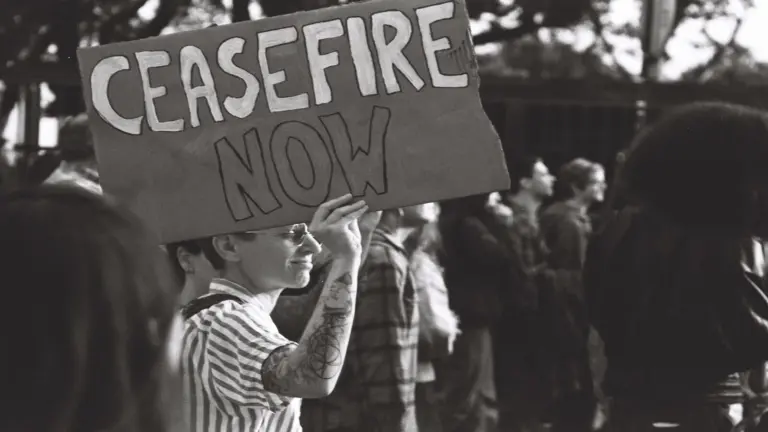 A person holds a "Ceasefire Now" sign at a protest. They're wearing glasses and a striped shirt, surrounded by others in a peaceful atmosphere.