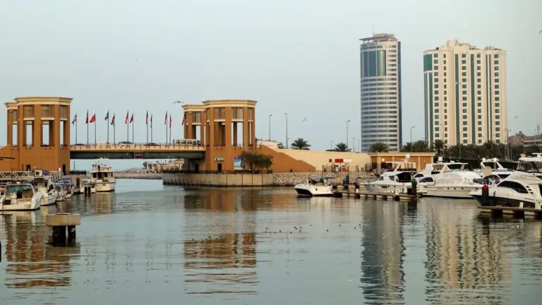 One of the strikes ended with a missile landing nearby United States 5th Fleet Headquarters in Manama, Bahrain, with a bridge adorned with flags spanning a calm marina. Modern high-rise buildings and moored yachts enhance the urban waterscape.