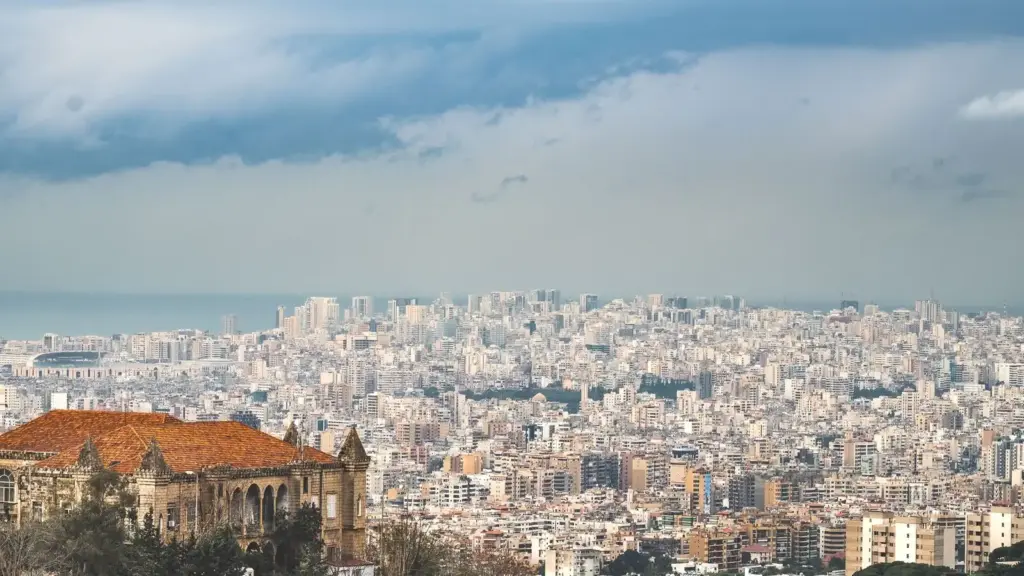 A sprawling cityscape of Beirut, Lebanon, under a cloudy sky, with dense urban buildings stretching to the horizon and a historic structure in the foreground.