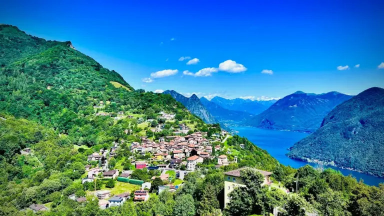 A lush green hillside village overlooks a serene blue lake with mountains in the background under a bright blue sky, evoking tranquility and natural beauty. Shows a beautiful landscape of Europe and European history.