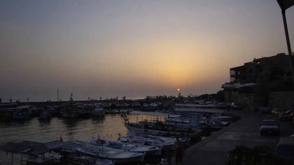Sunset over a tranquil harbor in Lebanon with moored boats lining the docks. Silhouetted figures walk along the distant horizon, and buildings overlook the scene.