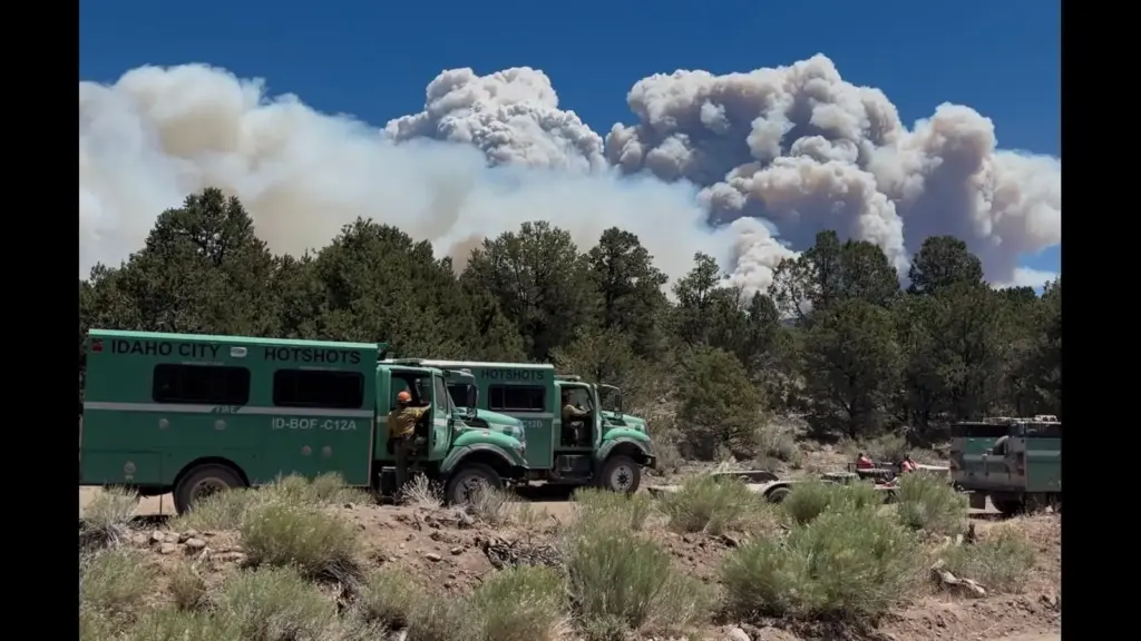 Two green fire trucks labeled "Idaho City Hotshots" are parked in a forest clearing, with smoke billowing in the background, indicating a wildfire.