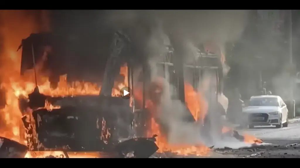 A thumbnail screenshot of a video from Belfast, Ireland showing a bus on fire at night, with flames rising from the front and thick gray smoke filling the scene.