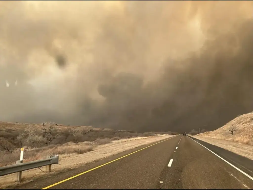 A wide-angle, low-resolution shot from a vehicle dashboard looking down a two-lane asphalt road under a severe, stormy sky. On the left side of the road, a dark, rotating funnel cloud extends from thick, low-hanging dark clouds toward the ground. The sky above is a heavy, hazy orange-brown hue, indicating a dusty or severe supercell thunderstorm environment. Snow patches or dry grass line the sides of the road.