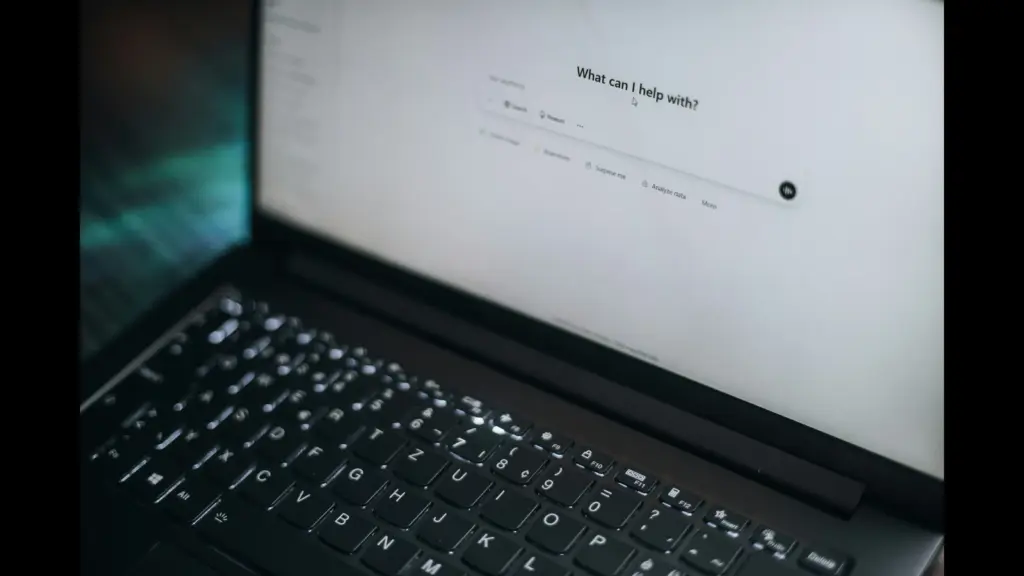 A black laptop sits open at an angle, showing its keyboard and a brightly lit white screen with blurred text and a single black dot or icon for ChatGPT and OpenAI. The background behind the laptop is dark and out of focus.