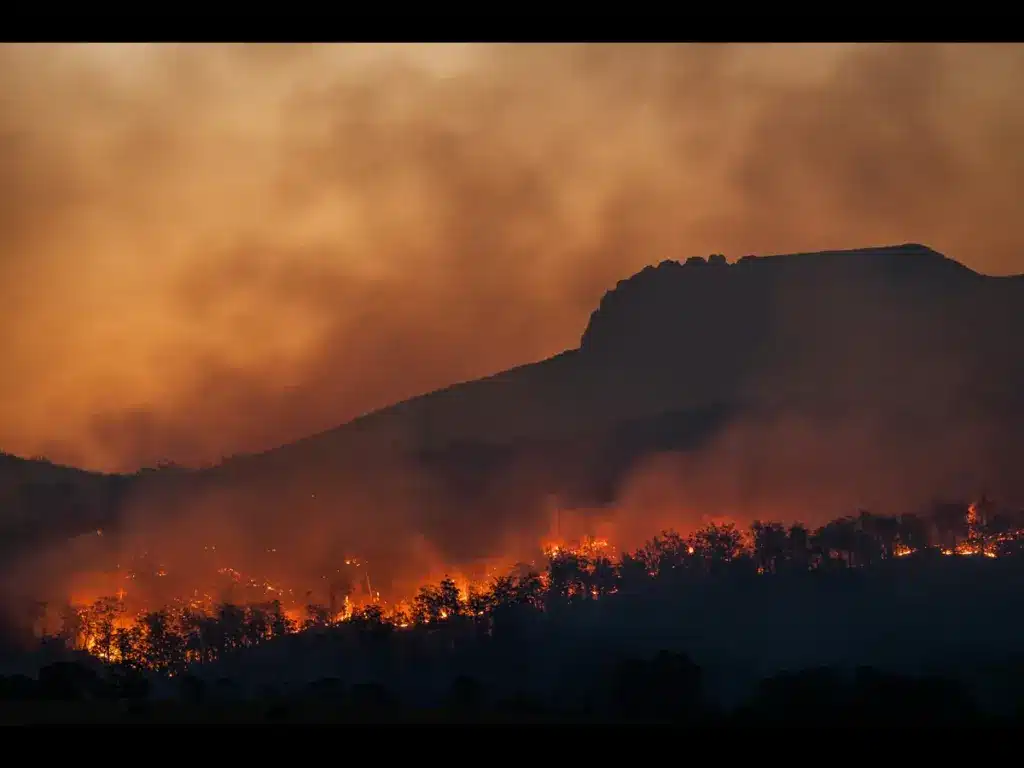 A large wildfire burning at twilight along the base of a dark, rocky mountain ridge. Thick, orange-and-brown smoke fills the sky above. Bright red and orange flames consume the silhouetted forest trees below.