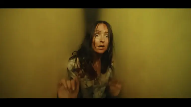 Frightened woman in the movie "Backrooms" with long dark hair raises her hands in a dim, yellow-lit hallway, staring ahead with a tense, alarmed expression.