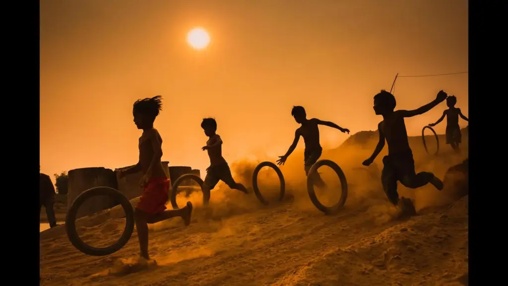 Silhouetted children run and play with rolling tires across a dusty field at sunset, creating a joyful, energetic scene in warm orange light.
