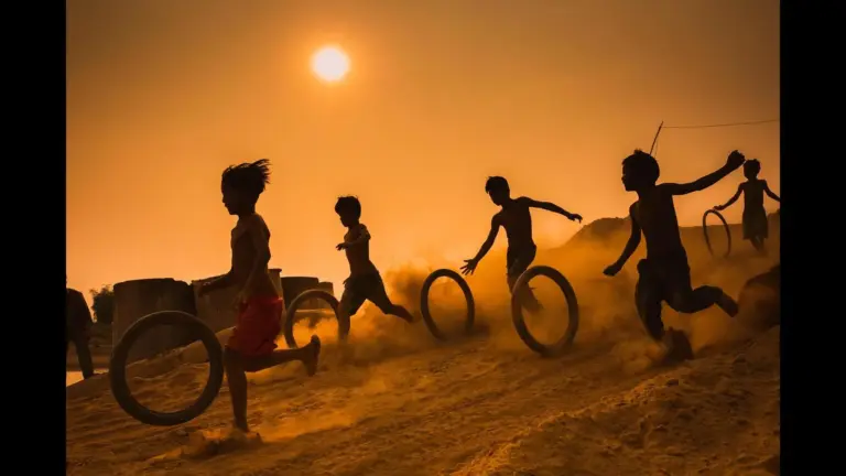 Silhouetted children run and play with rolling tires across a dusty field at sunset, creating a joyful, energetic scene in warm orange light.